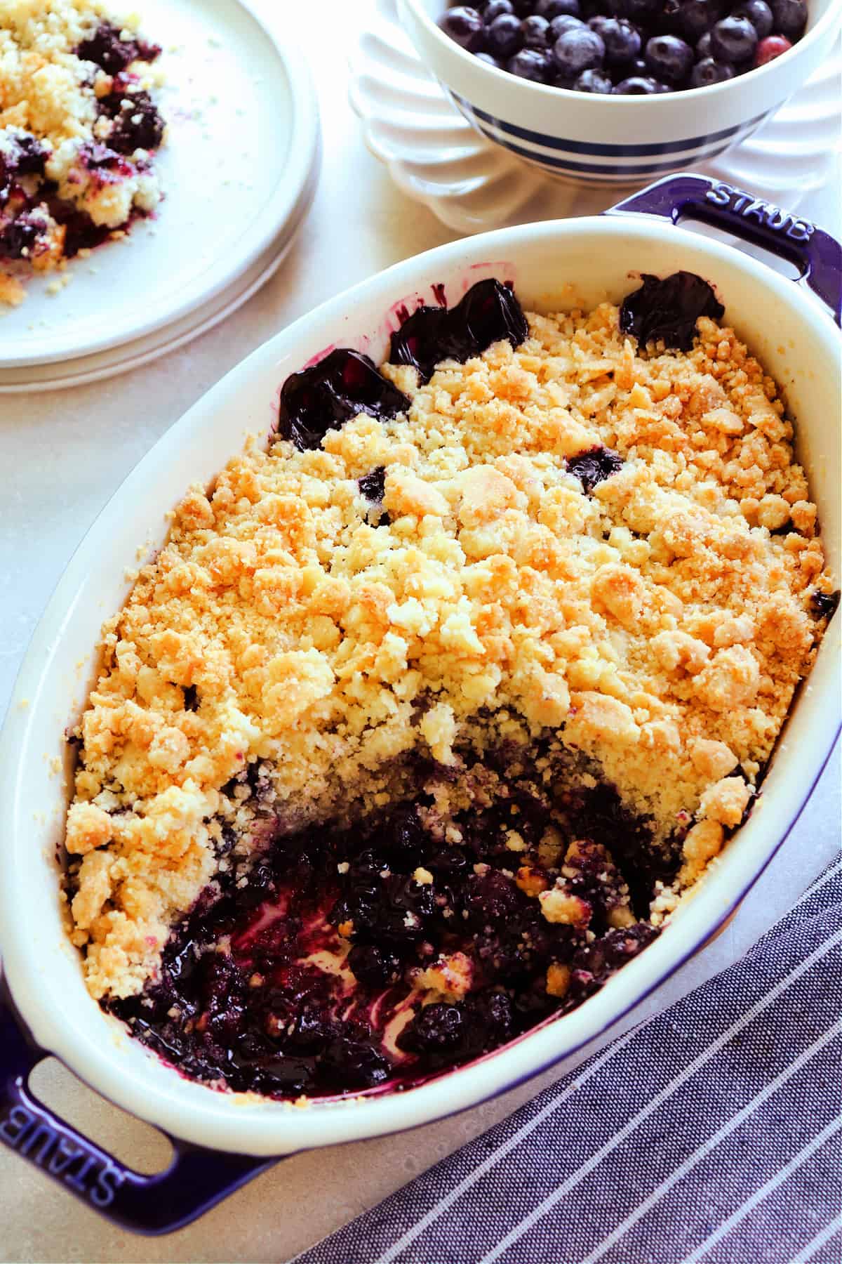 Blueberry crumble in a blue stoneware baking dish with a serving missing, with blue stripped towel next to it, and blueberries in a bowl behind it.