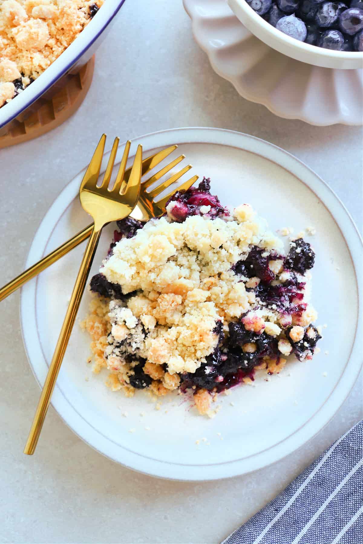 A serving of blueberry crumble on a small dessert plate, with two golden forks on the side.