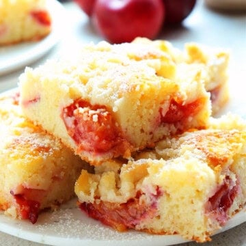 Close up photo of slices of coffee cake with plums on a white dessert plate, fresh plums in the back.