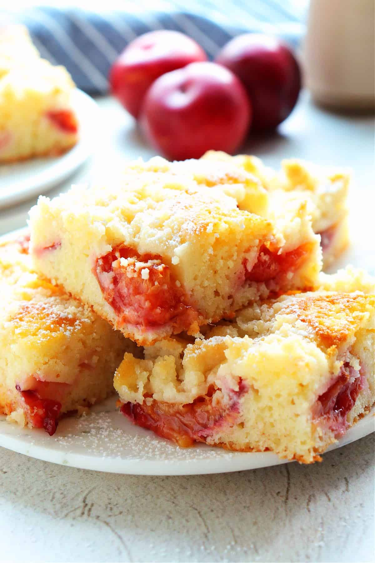 Close up photo of slices of coffee cake with plums on a white dessert plate, fresh plums in the back.