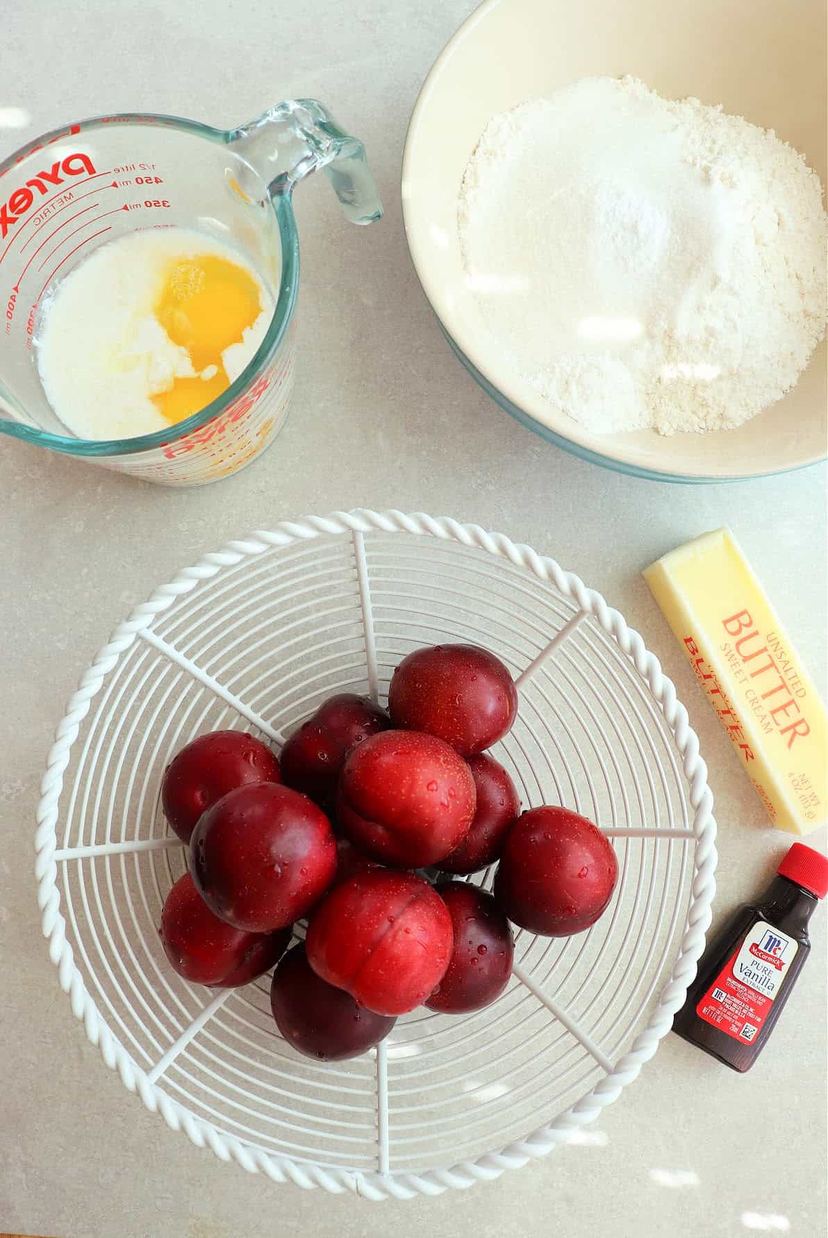 Dry cake ingredients in a bowl, a bottle of vanilla extract, stick of butter, buttermilk with eggs in a glass measuring cup and fresh plums in a white basket.